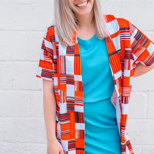 May include: A woman in a turquoise dress and a short-sleeved, open-front kimono. The kimono has a patchwork design with orange, white, and blue rectangular patterns. The woman is smiling, and the background is a white brick wall.