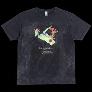 Puede incluir: Camiseta gris oscuro con una ilustración colorida de una rana arbórea, el texto "hang in there" y el logotipo de National Geographic. La rana es verde, roja y amarilla. La camiseta es de cuello redondo.