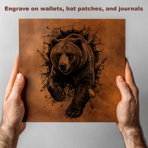 May include: A brown leather-like square with a detailed illustration of a bear breaking through a wall. The text "Engrave on wallets, hat patches, and journals" is at the top. The image is held by two hands.