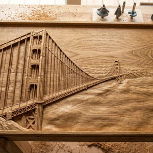 May include: A detailed wood carving of the Golden Gate Bridge in San Francisco. The bridge is depicted in a three-dimensional relief, showcasing its iconic towers and suspension cables. The artwork is set within a wooden frame.