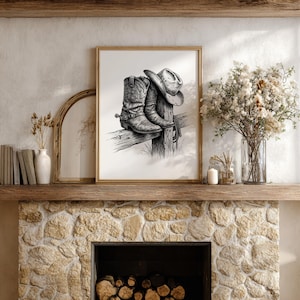 May include: A framed black and white print featuring a cowboy hat and boots resting on a wooden fence post. The artwork is displayed above a stone fireplace, with decorative elements like a vase of flowers and books.