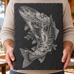 May include: A rectangular black slate plaque featuring a detailed white engraved illustration of a large fish. Inside the fish silhouette is a landscape scene showing a person fishing in a river, surrounded by pine trees and mountains.