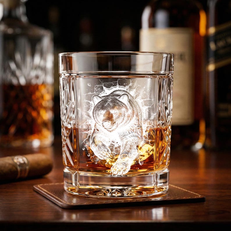 May include: A clear, square-shaped whiskey glass with a detailed etched image of a bear. The glass is filled with amber-colored liquid and sits on a brown leather coaster. The background includes bottles of liquor.