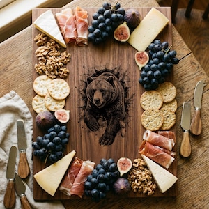 May include: A wooden charcuterie board featuring a detailed illustration of a bear breaking through a wall. The board is arranged with grapes, cheese, crackers, figs, walnuts, and prosciutto. Includes cheese knives.