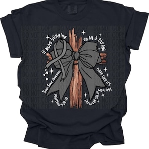 May include: Black t-shirt with a gray bow and wooden cross graphic. Text includes "If you're hanging on by a thread, make sure it's the hem of His garment." Also includes "Brain Tumor Awareness" and "Matthew 9:20-22."