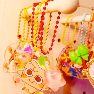 Puede incluir: Una colección de colgantes de acrílico transparente y colorido con varios diseños, incluyendo personajes de dibujos animados y alimentos, colgados de una barra de madera blanca. Los colgantes están unidos a collares de cuentas rojas, amarillas y otros colores.