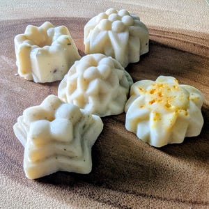 May include: An overhead shot of five handmade soaps in various shapes. The soaps are off-white with some having a speckled appearance. One is star-shaped, and others are floral or geometric. Some have a yellow dusting.