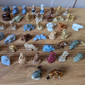 May include: A selection of miniature ceramic animal ornaments in shades of brown, blue, and white. The figurines represent creatures like cats, bears, and rabbits. The ornaments are displayed on a wooden surface.