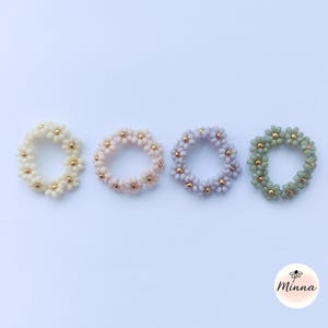 Könnte beinhalten: Vier ringförmige Schmuckstücke in Form von Gänseblümchen in Pastellfarben: Creme, Pfirsich, Lavendel und Salbeigrün. Jeder Ring hat kleine, runde Perlen mit goldfarbenen Zentren. Die Ringe liegen auf einer weißen Oberfläche.