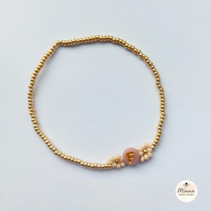 Könnte beinhalten: Goldfarbenes Perlenarmband mit einer rosa Perle mit dem Buchstaben "F" und kleinen Blumenakzenten. Das Armband besteht aus kleinen, runden Goldperlen und ist als Accessoire konzipiert.