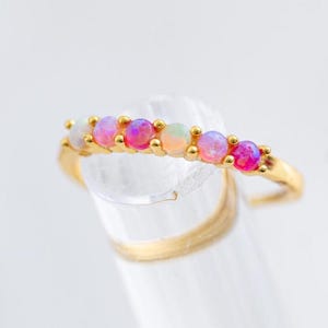 May include: A gold ring featuring a row of colorful opal gemstones. The opals are in shades of pink, purple, and white. The ring is set on a clear, cylindrical object, with a gold band visible.