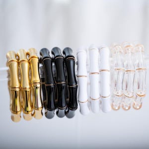 May include: A collection of bamboo-style bracelets in gold, black, white, and clear. Each bracelet features a repeating pattern of segmented cylinders, with gold accents on the gold, black, and clear bracelets. The white bracelets have gold accents.