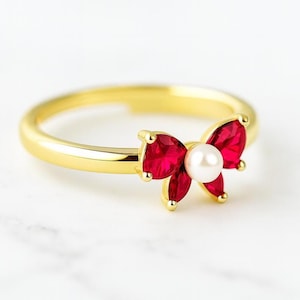 May include: A gold-colored ring featuring a bow design with red gemstones and a central white pearl. The ring's band is smooth and polished, and the bow is the focal point of the jewelry piece.