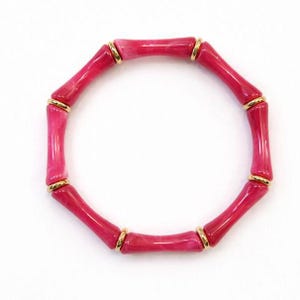 May include: A pink bamboo-style bracelet with gold-colored accents. The bracelet is made of several connected segments, each a vibrant shade of pink. The gold accents are circular and connect the pink segments.
