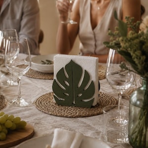 Pode incluir: Um porta-guardanapos verde-escuro em forma de folha de monstera, contendo uma pilha de guardanapos de papel quadrados brancos. O suporte está posicionado sobre um lugar americano circular trançado numa mesa com copos, pratos e uvas verdes.