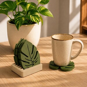 Pode incluir: Um conjunto de bases para copos verde-escuras em forma de folha com um suporte retangular a condizer, e uma caneca de cerâmica bege salpicada pousada sobre uma base individual. As bases apresentam um design recortado detalhado de folha.