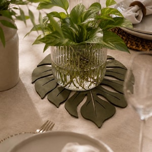 Peut inclure: Un set de table vert foncé en forme de feuille, fabriqué dans une matière lisse, placé sous un vase en verre transparent à côtes verticales contenant une plante verte au feuillage panaché. Le set présente des découpes imitant les nervures.