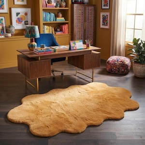 Könnte beinhalten: Ein großer, unregelmäßig geformter, wolkenartiger Teppich in einfarbigem Senfgelb mit einer weichen, flauschigen Kunstfelltextur. Der Teppich liegt auf einem dunklen Holzboden vor einem Schreibtisch und setzt einen strukturierten Akzent.