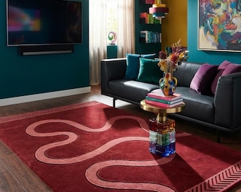May include: A rectangular area rug in deep red featuring a lighter red, winding, serpentine line pattern across the center and a textured, chevron-style border design at both ends, laid flat on a dark wood floor in a living room setting.