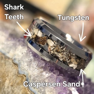 May include: A tungsten ring with a band of Caspersen sand and inlaid with shark teeth. The ring is silver in color and sits on a purple and white surface.