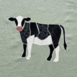 May include: A sage green fabric background featuring a cow applique. The cow is constructed from black floral patterned fabric, solid black fabric, and white fabric pieces, all outlined with dark stitching. It has a small pink nose detail.
