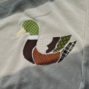 May include: A beige crewneck sweatshirt featuring a large, central appliqué of a mallard duck. The duck is constructed from various fabric patches in green, brown, tan, and plaid patterns, with visible stitching outlining each section of the design.