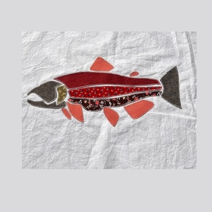 Peut inclure: Un textile en tissu blanc présentant une applique de poisson centrale. Le poisson est composé de divers morceaux de tissu dans des tons rouge foncé, marron et corail, avec des motifs floraux sur certaines sections et une tête et une queue marron foncé.