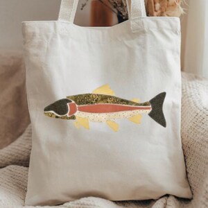 May include: A cream-colored canvas tote bag featuring a central graphic of a fish. The fish design is composed of various fabric patches in shades of olive green, dark charcoal, muted red, and pale yellow, showcasing a speckled pattern.