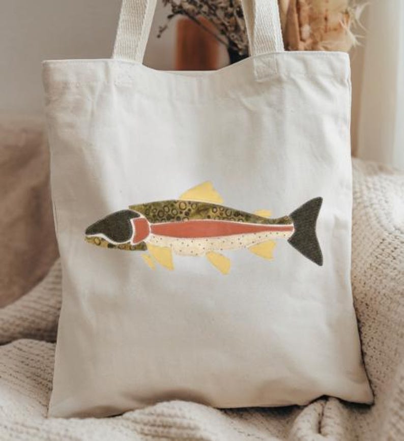 May include: A cream-colored canvas tote bag featuring a central graphic of a fish. The fish design is composed of various fabric patches in shades of olive green, dark charcoal, muted red, and pale yellow, showcasing a speckled pattern.