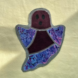 Op de afbeelding: Een stoffen applicatie in de vorm van een geest met een donkerpaarse kop en romp, voorzien van zwarte ronde ogen en een mond. De geest heeft vleugels en een onderkant van paars en blauw patroonstof, afgewerkt met een grijze zigzagsteek.