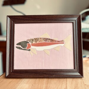 May include: A framed artwork featuring a stylized fish collage on a light pink fabric background. The fish is composed of various patterned and solid-colored fabric segments, including leopard print, solid rust, and cream, all encased in a dark brown frame.