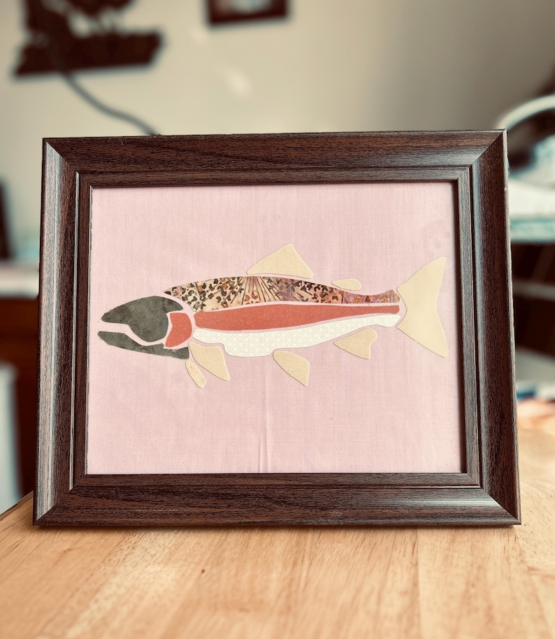 May include: A framed artwork featuring a stylized fish collage on a light pink fabric background. The fish is composed of various patterned and solid-colored fabric segments, including leopard print, solid rust, and cream, all encased in a dark brown frame.