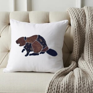 May include: A square white throw pillow featuring a central applique design of a beaver. The beaver is constructed from various fabric patches in shades of brown and dark blue with a subtle pattern, outlined with visible decorative stitching.