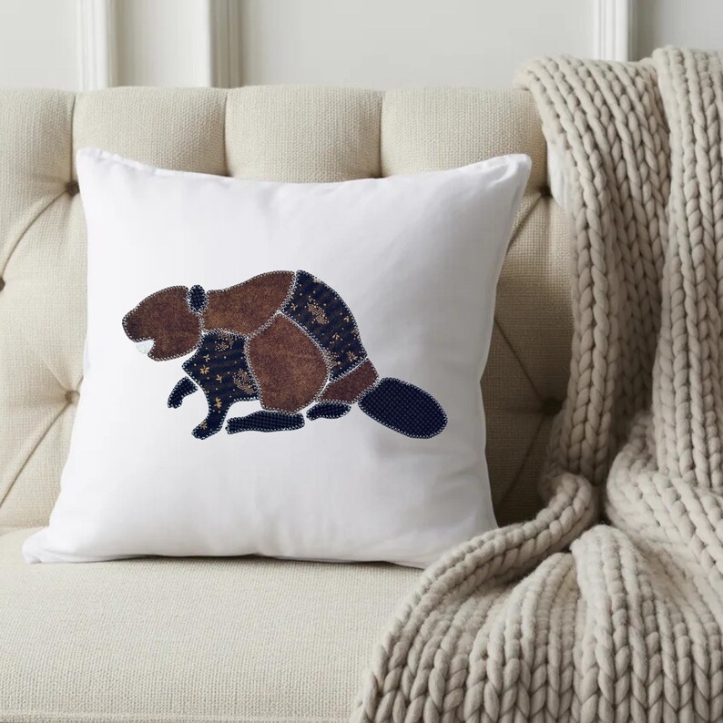 May include: A square white throw pillow featuring a central applique design of a beaver. The beaver is constructed from various fabric patches in shades of brown and dark blue with a subtle pattern, outlined with visible decorative stitching.