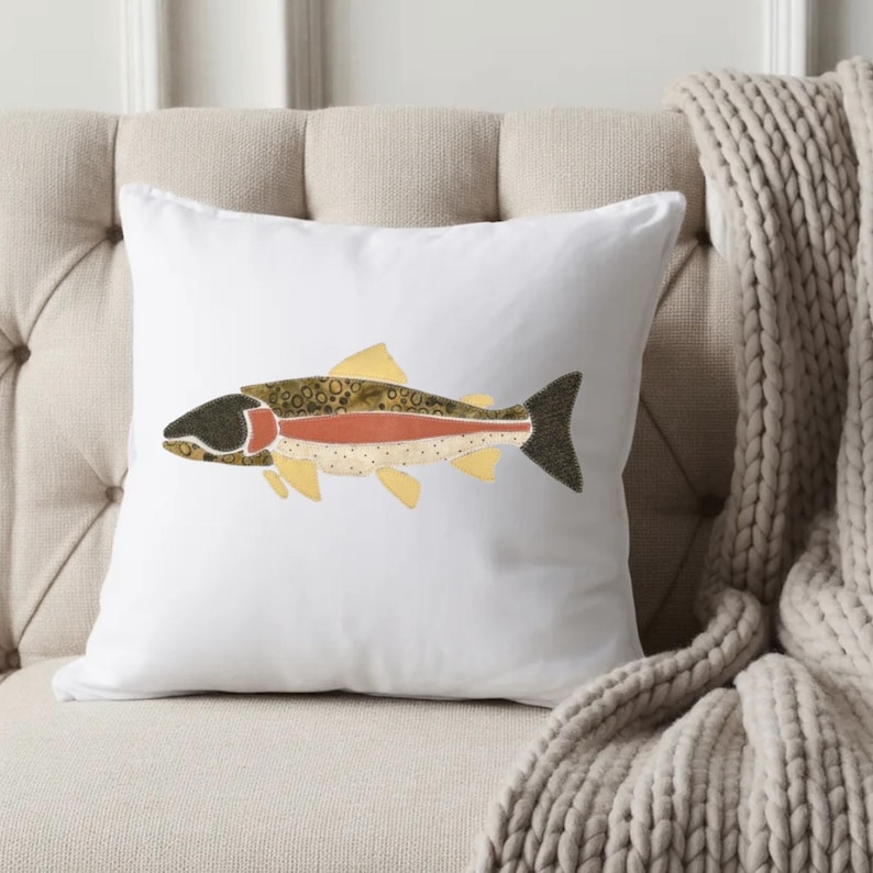 May include: A square white throw pillow featuring a centered graphic illustration of a fish. The fish design incorporates textured patterns in shades of olive green, dark brown, cream, and a prominent horizontal stripe of muted terracotta orange.
