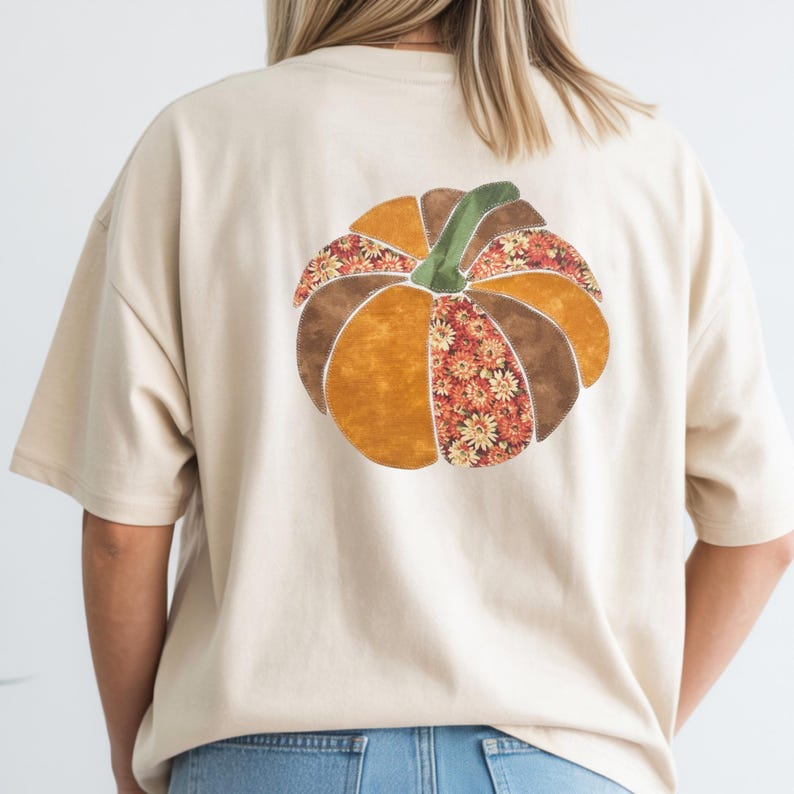 May include: A cream-colored short-sleeve t-shirt featuring a large pumpkin graphic on the back. The pumpkin is composed of various fabric-like segments in shades of orange, brown, and green, with some sections displaying a floral pattern.