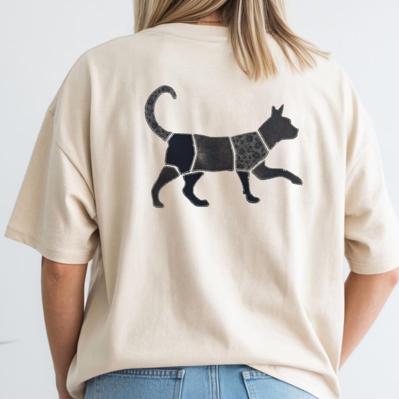 May include: A beige short-sleeved t-shirt featuring a graphic on the back of a cat silhouette. The cat is composed of several distinct dark-colored segments with varied textures and patterns, outlined with a visible white stitched border design.