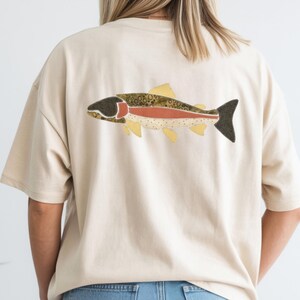 May include: A cream-colored short-sleeve t-shirt featuring a large graphic on the back. The graphic depicts a stylized fish with a dark head, a reddish-orange horizontal stripe along its body, and a textured pattern of brown spots and yellow fins.