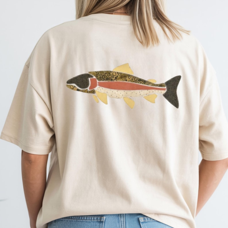 May include: A cream-colored short-sleeve t-shirt featuring a large graphic on the back. The graphic depicts a stylized fish with a dark head, a reddish-orange horizontal stripe along its body, and a textured pattern of brown spots and yellow fins.