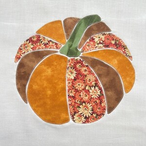 May include: A fabric applique of a pumpkin centered on a white background. The pumpkin is constructed from segments of solid mustard yellow, solid brown, and floral patterned fabric in shades of orange, yellow, and brown, with a green stem.