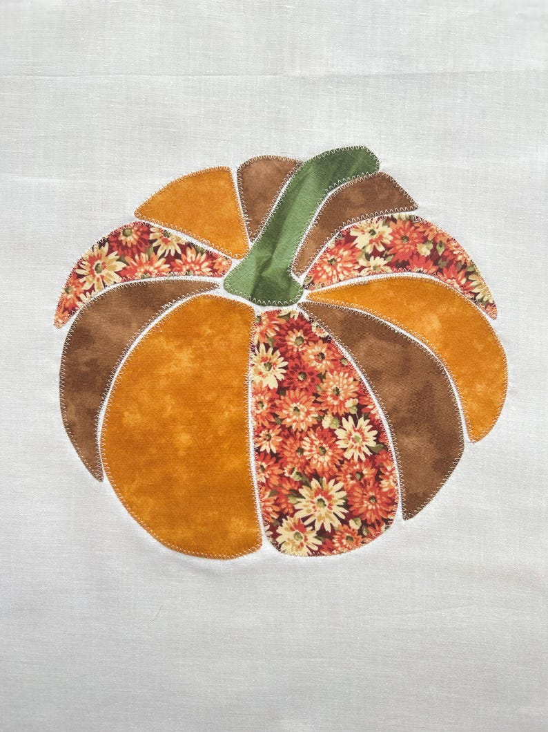 May include: A fabric applique of a pumpkin centered on a white background. The pumpkin is constructed from segments of solid mustard yellow, solid brown, and floral patterned fabric in shades of orange, yellow, and brown, with a green stem.