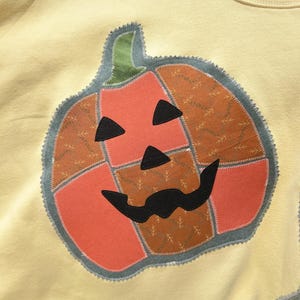 May include: A pale yellow sweatshirt featuring a large, central pumpkin appliqu&eacute;. The pumpkin is constructed from patchwork fabric sections in orange and brown with a leaf pattern, accented by black fabric eyes, a nose, and a moustache.
