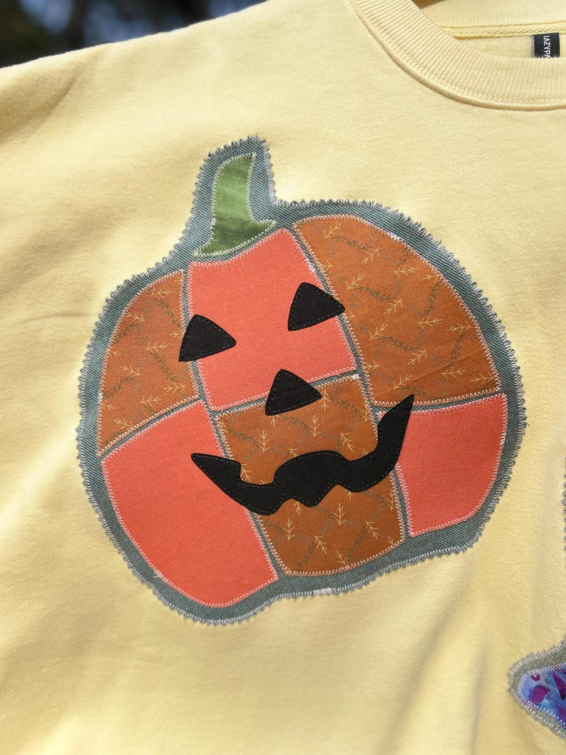 May include: A pale yellow sweatshirt featuring a large, central pumpkin appliqu&eacute;. The pumpkin is constructed from patchwork fabric sections in orange and brown with a leaf pattern, accented by black fabric eyes, a nose, and a moustache.