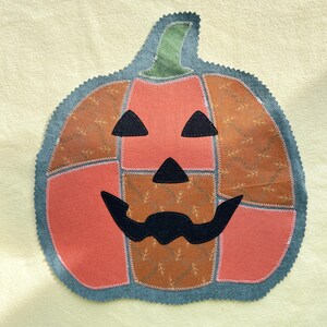 May include: A fabric pumpkin appliqu&eacute; featuring a patchwork design of solid coral and brown patterned fabric sections. The jack-o'-lantern face consists of black fabric eyes, a nose, and a wide mouth, all outlined with white stitching.