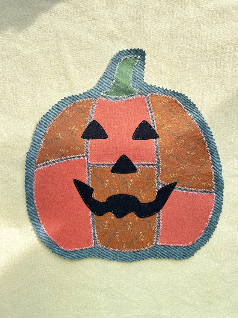 May include: A fabric pumpkin appliqu&eacute; featuring a patchwork design of solid coral and brown patterned fabric sections. The jack-o'-lantern face consists of black fabric eyes, a nose, and a wide mouth, all outlined with white stitching.