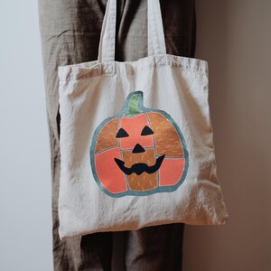 May include: A beige canvas tote bag featuring a large, segmented pumpkin graphic. The pumpkin is composed of orange and brown patterned fabric patches with a dark teal outline, black triangular eyes, a black nose, and a black mouth.