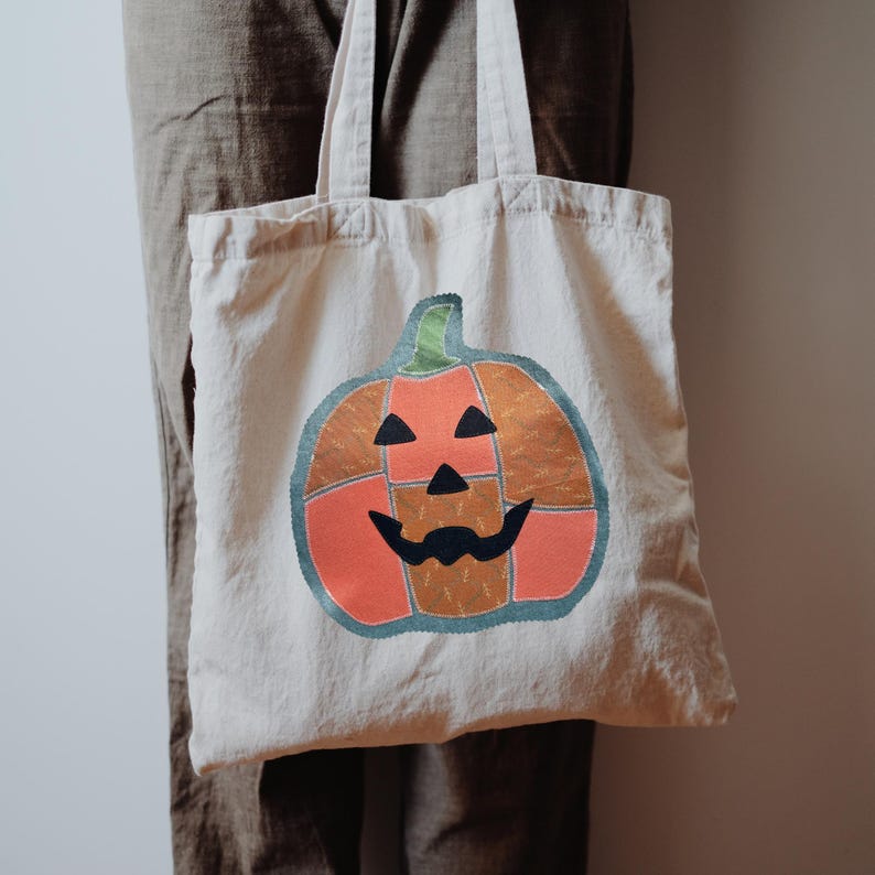 May include: A beige canvas tote bag featuring a large, segmented pumpkin graphic. The pumpkin is composed of orange and brown patterned fabric patches with a dark teal outline, black triangular eyes, a black nose, and a black mouth.