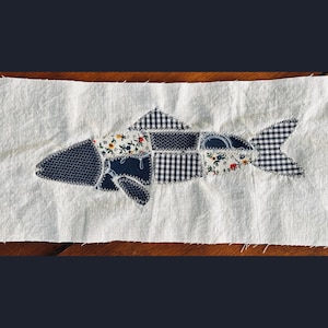 May include: A rectangular piece of off-white fabric featuring a fish-shaped appliqué made from various fabric scraps. The fish is constructed from navy blue, white, and black patterned textiles, including gingham, floral, and geometric prints.