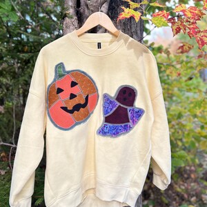 Op de afbeelding: Een lichtgeel sweatshirt met ronde hals en twee grote stoffen applicaties op de voorzijde. E&eacute;n applicatie is een oranje pompoen met een zwart gezicht, de andere is een spookje van donkerpaarse en blauwgepatroneerde stof met een zwart gezicht.