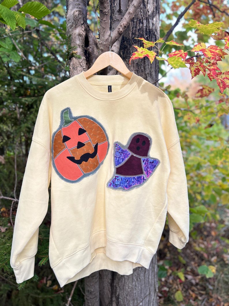 Op de afbeelding: Een lichtgeel sweatshirt met ronde hals en twee grote stoffen applicaties op de voorzijde. E&eacute;n applicatie is een oranje pompoen met een zwart gezicht, de andere is een spookje van donkerpaarse en blauwgepatroneerde stof met een zwart gezicht.